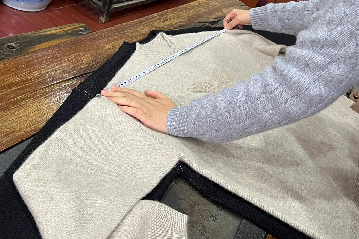 Hands checking the width of a light-colored knit sweater with a measuring tape.