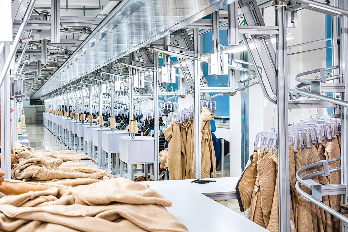 Knitted garments hanging along overhead rails beside rows of factory workstations.