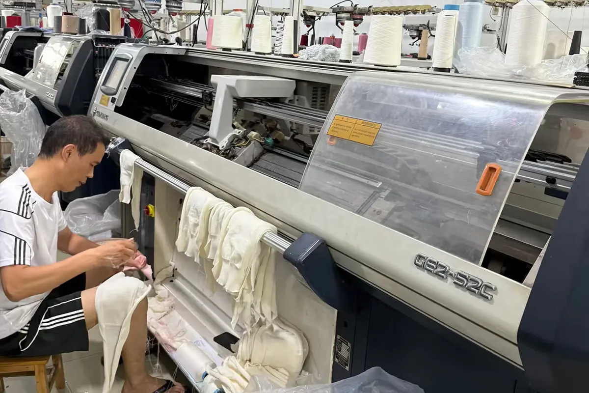 Worker handling knitted pieces beside a flat knitting machine.