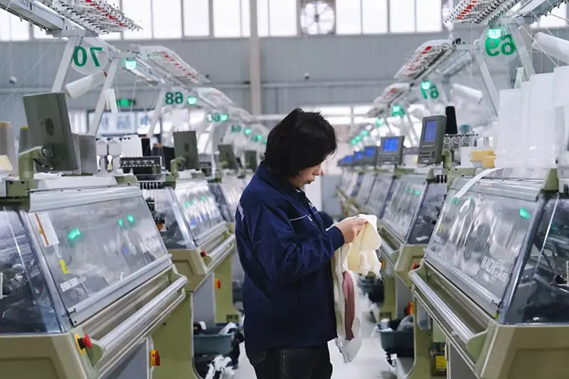 Worker inspecting knitted fabric between rows of computerized knitting machines.