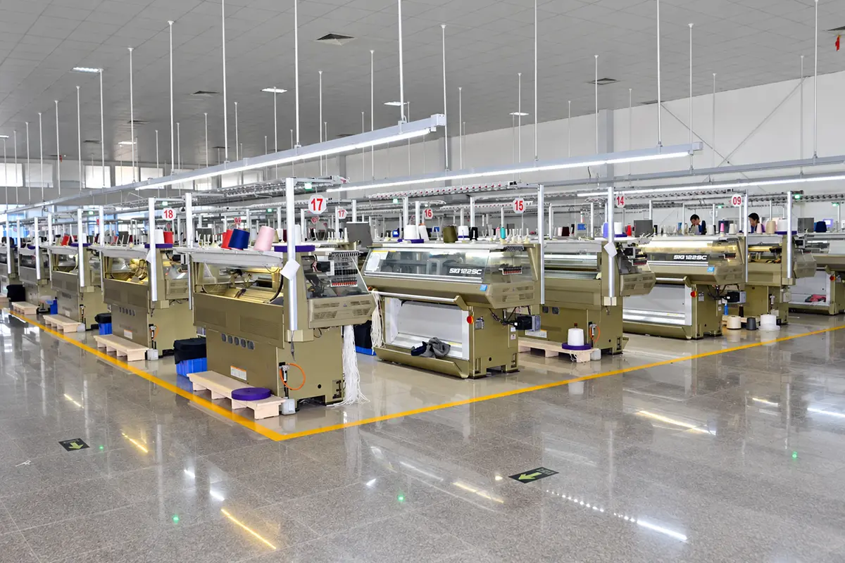 Rows of flat knitting machines and yarn cones in a spacious factory workshop.